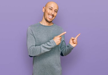 Sakallı, sıradan kıyafetler giyen, gülümseyen ve kameraya bakan kel bir adam. İki eliyle ve parmaklarıyla kenarı işaret ediyor.. 