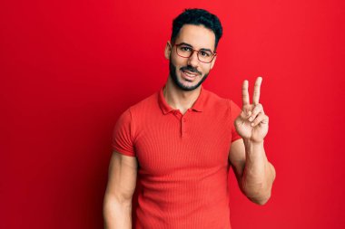 Gündelik kıyafetler giyen ve gözlüklü genç İspanyol adam iki numarayı gösterip işaret ederken kendinden emin ve mutlu bir şekilde gülümsüyor.. 