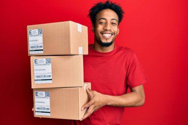 Sakallı, teslimat paketleri taşıyan, yüzünde mutlu ve havalı bir gülümsemeyle gülümseyen genç Afrikalı Amerikalı bir adam. diş gösterme. 