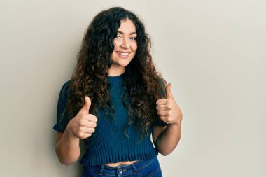 Kıvırcık saçlı, sıradan giyinen genç esmer kadın. Başarı işareti yapıyor. Elleriyle olumlu jest yapıyor. Neşeli ifade ve kazanan jest. 