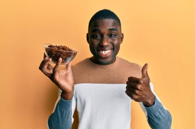 Genç Afro-Amerikalı elinde kuru üzümlü bir kaseyle mutlu ve pozitif gülümsüyor, başparmağıyla mükemmel ve onay işareti yapıyor. 