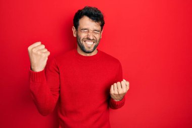 Sakallı, sıradan kırmızı kazak giyen yakışıklı adam kolları havada ve gözleri kapalı olarak başarıyı kutluyor ve şaşırıyor. Kazanan konsept. 