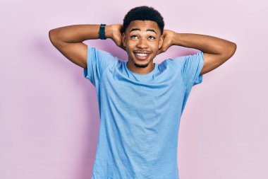 Genç Afro-Amerikalı adam günlük mavi tişört giyiyor rahatlıyor ve esniyor, kolları ve elleri başının arkasında mutlu bir şekilde gülümsüyor. 