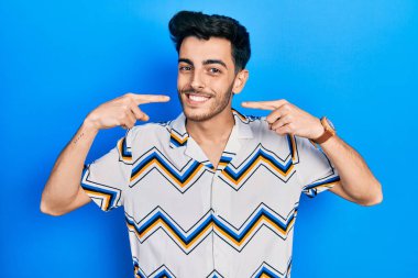 Günlük kıyafetler giyen, neşeli gülümseyen, parmaklarıyla ve ağzıyla işaret eden İspanyol genç bir adam. diş sağlığı kavramı. 