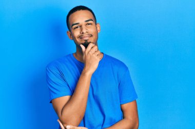 Sıradan mavi tişörtler giyen genç Afro-Amerikan bir adam kameraya kendinden emin bakıyor kollarını kavuşturmuş ve çenesini kaldırmış gülümsüyor. Olumlu düşünmek. 