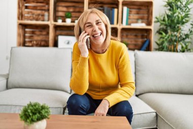 Orta yaşlı sarışın kadın akıllı telefonda konuşuyor. Evdeki kanepede oturuyor..