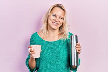 Kafkasyalı güzel sarışın kadın bir fincan kahve içiyor. Elinde termo var ve kameraya seksi bir ifadeyle bakıyor. Neşeli ve mutlu bir yüzü var.. 