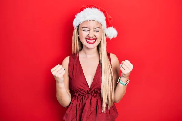 年轻的金发姑娘戴着圣诞礼帽 非常高兴而兴奋地举起双臂 做着获胜的手势 微笑着 尖叫着要成功 庆祝概念 — 图库照片