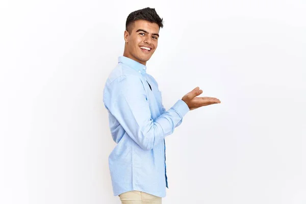 ビジネスシャツを身に着けている若いヒスパニック系の男は コピースペースを示す手を開いて手のひらで脇を指して孤立した背景の上に立って 広告笑顔興奮幸せ — ストック写真