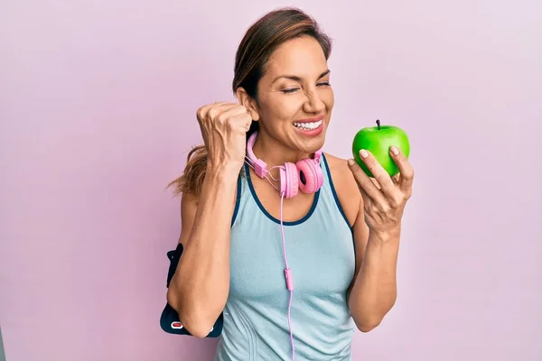 年轻的拉丁女人穿着运动服 用耳机 吃着绿色苹果 自豪地尖叫着 高举双臂庆祝胜利和成功 — 图库照片
