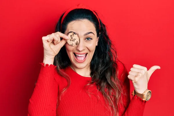 Young Hispanic Woman Holding Cookie Eye Pointing Thumb Side Smiling — Stock Photo, Image