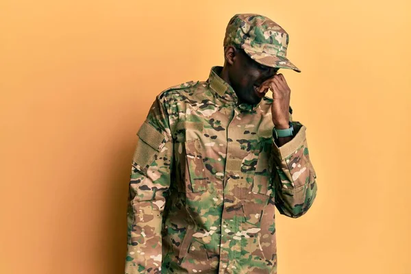 Young african american man wearing army uniform smelling something stinky and disgusting, intolerable smell, holding breath with fingers on nose. bad smell
