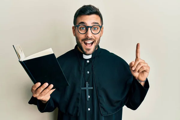 Joven Sacerdote Hispano Sosteniendo Biblia Con Dedo Alto Sonriendo Riendo —  Fotos de Stock