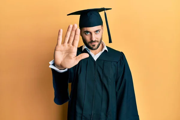 卒業キャップと儀式用のローブを着た若いヒスパニック系の男が手の手のひらで歌うのをやめます 顔に負の重大なジェスチャーで — ストック写真