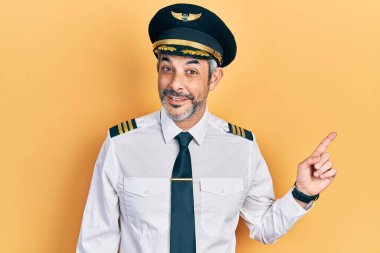 Yakışıklı, orta yaşlı, gri saçlı, uçak pilotu üniforması giyen, yüzünde kocaman bir gülümseme olan, eli kameraya bakan bir adam.. 