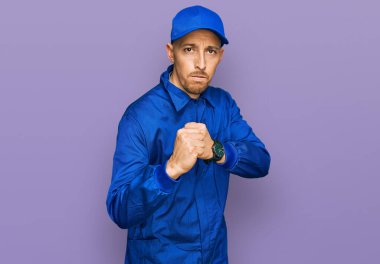 Kel ve sakallı bir adam, inşaatçı tulumu giyiyor. Yumruk savunma hareketiyle dövüşmeye hazır. Kızgın ve üzgün. 