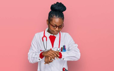Doktor üniforması ve steteskop giyen genç Afrikalı Amerikalı kadın kol saatinde saati kontrol ediyor, rahat ve kendinden emin. 