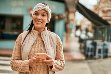 Genç Latin kadın şehirde akıllı telefon kullanarak mutlu bir şekilde gülümsüyor..