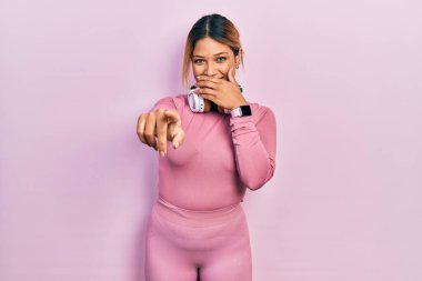 Güzel İspanyol kadın spor kıyafetleri giyiyor ve kulaklık takıyor. Sana gülüyor, eli ağzında kamerayı işaret ediyor. Utanç ifadesi. 