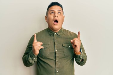 Gündelik kıyafetler giyen genç latin adam şaşırdı ve şaşırdı. Yukarı baktı ve parmaklarıyla işaret etti ve kollarını kaldırdı.. 