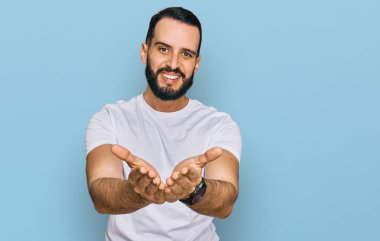 Sakallı, sıradan beyaz tişörtlü, elleri avuçlarında gülümseyen ya da el hareketi yapan genç bir adam. Bekleme ve koruma 