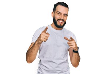 Sakallı, sıradan beyaz tişörtlü, mutlu ve komik yüzlü genç adam kamerayı işaret ediyor. iyi enerji ve titreşimler. 