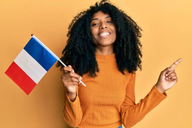 Afro saçlı Afrika kökenli Amerikalı kadın Fransa bayrağı tutarken mutlu bir şekilde gülümsüyor. 