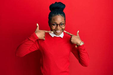 Genç Afro-Amerikalı kadın günlük kıyafetler ve gözlük takıyor neşeli bir şekilde gülümsüyor ve parmaklarıyla ve ağzıyla işaret ediyor. diş sağlığı kavramı. 