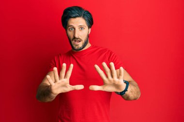 Günlük kırmızı tişört giyen genç İspanyol adam korku ifadesinden korkmuş ve dehşete düşmüş. El kol hareketi yapmayı kes, şok içinde bağırmayı kes. panik konsepti. 