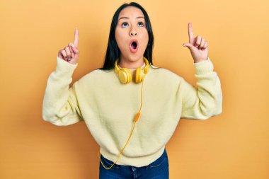 Boynuna kulaklık takan güzel İspanyol kadın şaşırmış ve şaşırmış bir şekilde yukarı bakıyor ve parmaklarıyla işaret ediyor ve kollarını kaldırıyor.. 