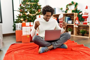 Genç Afro-Amerikalı adam dizüstü bilgisayarını kullanarak mutlu ve heyecanlı bir şekilde kollarını kaldırıp, gülümseyerek ve başarı çığlıkları atarak zafer jesti yapıyor. kutlama konsepti. 