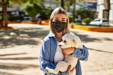 Koronavirüs maskesi takan genç sarışın kız şehirde köpeği tutuyor..