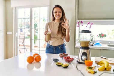 Güzel, genç, esmer bir kadın mutfakta bir bardak smoothie içiyor. Parmaklarıyla imza atıyor, gülümseyerek mükemmel bir sembol oluşturuyor. 