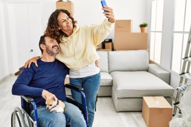 Orta yaşlı İspanyol çift mutlu gülümsüyor. Tekerlekli sandalyede oturan bir adam, bacaklarında köpek ve yeni evinde akıllı telefondan selfie çeken bir kadın..