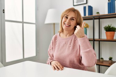 Orta yaşlı beyaz kadın, günlük giysiler içinde mutlu ve olumlu bir şekilde gülümseyerek evde oturuyor, başparmağıyla mükemmel ve onay işareti yapıyor. 