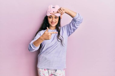 Ortadoğulu güzel bir kadın uyku maskesi ve pijama giyip gülümsüyor. Elleriyle ve parmaklarıyla gülen yüzüyle çerçeve yapıyor. yaratıcılık ve fotoğrafçılık kavramı. 