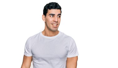 Yakışıklı, beyaz tişört giyen, yüzünde gülümseme ve doğal ifadeyle yan yana bakan bir İspanyol. Kendinden emin gülen. 