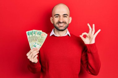 Arjantin Peso banknotları tutan genç kel adam parmaklarıyla imza atıyor, gülümseyerek mükemmel bir sembol oluşturuyor. 