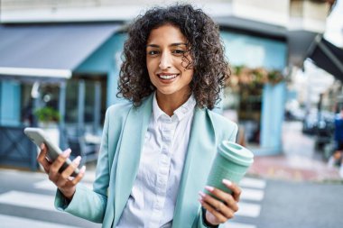 Profesyonel giyinen genç İspanyol iş kadını akıllı telefon kullanarak şehre gülümsüyor.