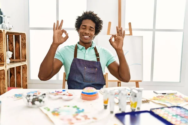 若いアフリカ系アメリカ人の男性がアートスタジオのテーブルの上に座って 指で瞑想のジェスチャーをして目を閉じてリラックスして笑顔 ヨガのコンセプト — ストック写真