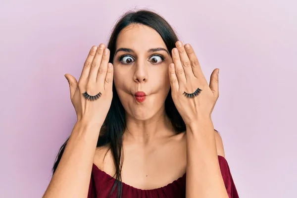 Young hispanic woman covering eyes with hands and fake lashes making fish face with mouth and squinting eyes, crazy and comical.