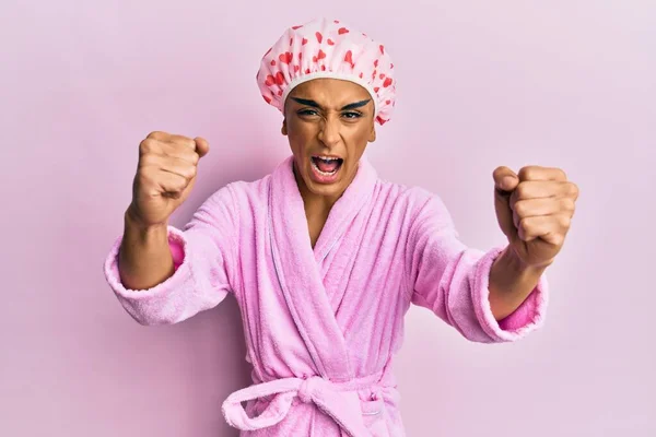 Hispanic man wearing make up wearing shower towel cap and bathrobe angry and mad raising fists frustrated and furious while shouting with anger. rage and aggressive concept.