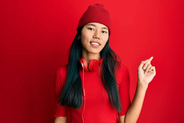 Young chinese woman listening to music using headphones with a big smile on face, pointing with hand and finger to the side looking at the camera.