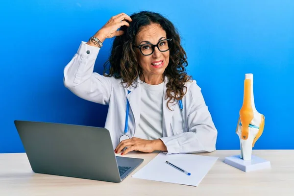 Beautiful middle age woman doctor at orthopedic clinic confuse and wonder about question. uncertain with doubt, thinking with hand on head. pensive concept.