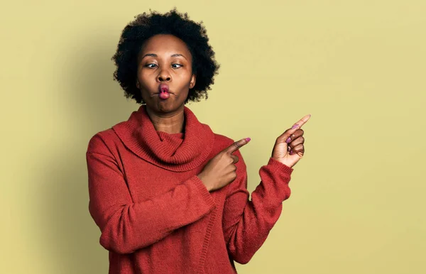 African american woman with afro hair pointing with fingers to the side making fish face with mouth and squinting eyes, crazy and comical.