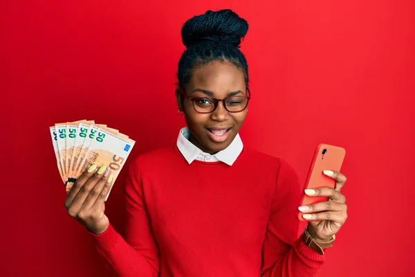 Joven Mujer Afroamericana Usando Smartphone Con Billetes Euros Guiñando Ojo —  Fotos de Stock