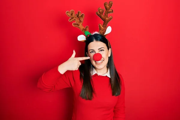 顔と鼻に手指で指差す鹿のクリスマスの帽子と赤い鼻を身に着けている若いヒスパニック系の女性は 陽気に笑っています 美の概念 — ストック写真