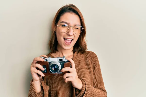 Young caucasian woman holding vintage camera winking looking at the camera with sexy expression, cheerful and happy face.