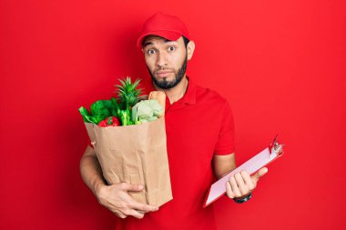 Hispanic man with beard wearing courier uniform with groceries from supermarket and clipboard clueless and confused expression. doubt concept. 
