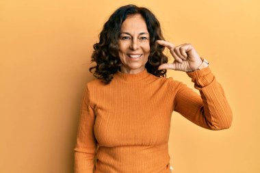 Orta yaşlı İspanyol kadın günlük kıyafetler giyiyor gülümsüyor ve el hareketleriyle küçük boy işaretler yapıyor parmaklarıyla ve kamerayla bakıyor. ölçüm kavramı. 